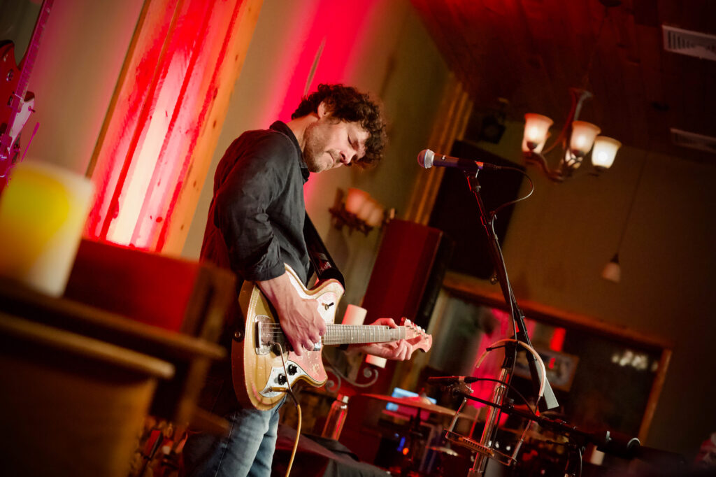 Musician playing electric guitar on stage, illuminated by red lights. He is focused and immersed, with a microphone nearby. The setting is warm and intimate.