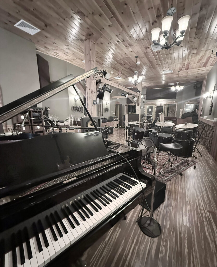 Wooden music studio with a grand piano and drum set under warm lighting. Cozy atmosphere with a rustic ceiling, creating a creative and inviting space.