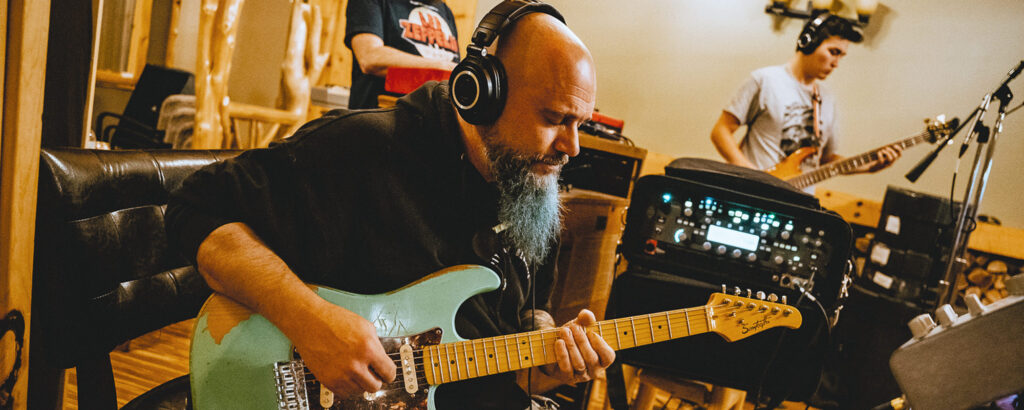 A bald musician with a blue beard focusedly plays an electric guitar, wearing headphones. Two other musicians play in the background, creating a collaborative, creative atmosphere.