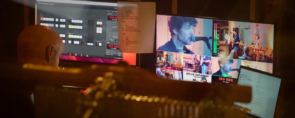 A person in a control room looks at live video feeds on screens. Monitors display a musician singing and performing. Warm lighting sets a focused tone.