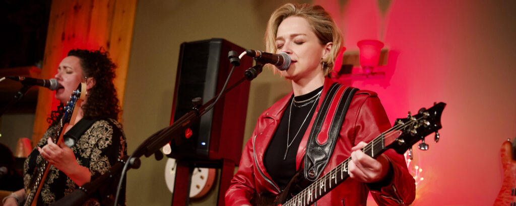 Two performers sing and play guitars on stage, illuminated by red lighting. The woman in a red leather jacket, Margo Macero takes center stage, conveying passion and energy.