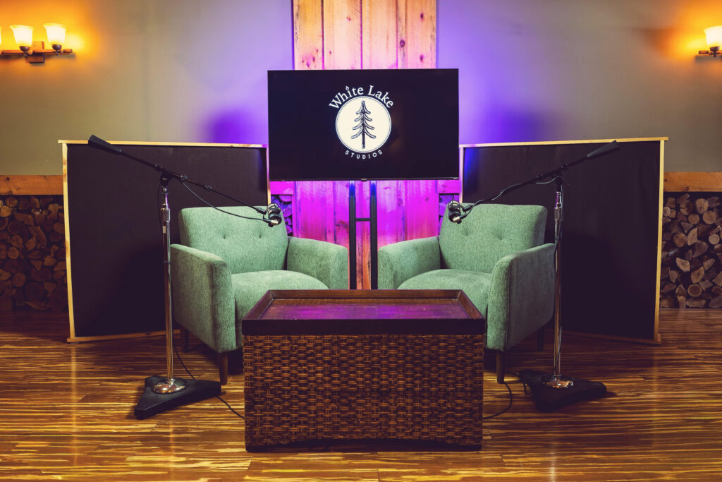 Cozy recording studio with two teal armchairs, microphones, and a wicker coffee table. A screen displays “White Lake Studios” on a warm wood backdrop.