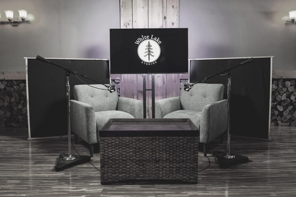 Two gray armchairs face each other with microphones on stands beside them, centered in front of a screen displaying "White Lake Studios," creating a cozy podcast or interview setting.
