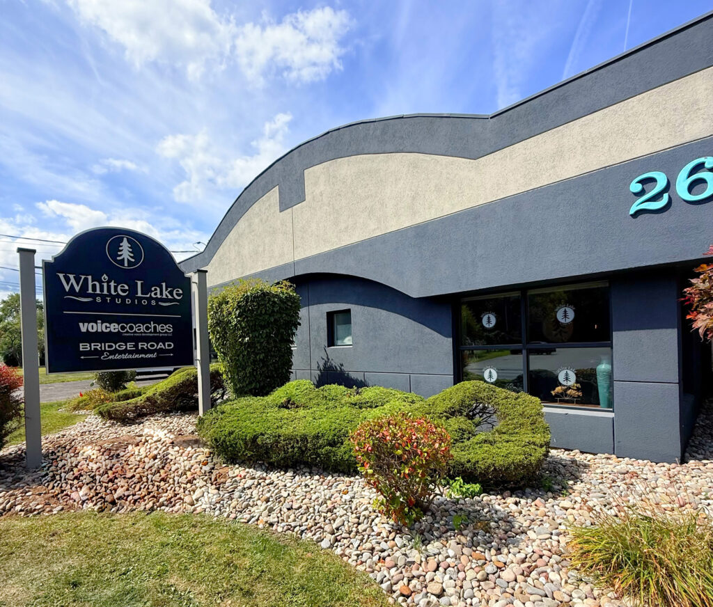 Exterior of a modern building with a curved roof, manicured shrubs, and a sign reading "White Lake Studios." Bright, sunny day with blue skies.